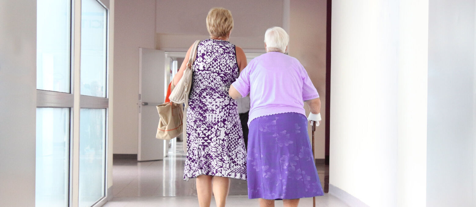 Ältere Dame geht eingehakt bei einer Angehörigen im Arm auf einem Klinikflur.
