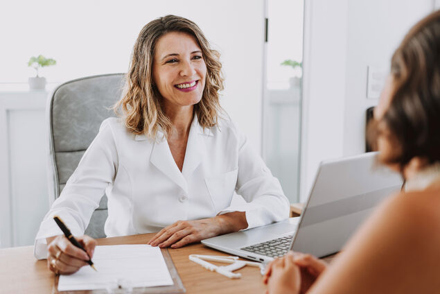 Frau mit blonden, lockigen Haaren füllt einen Fragenbogen mit einer Patientin aus.