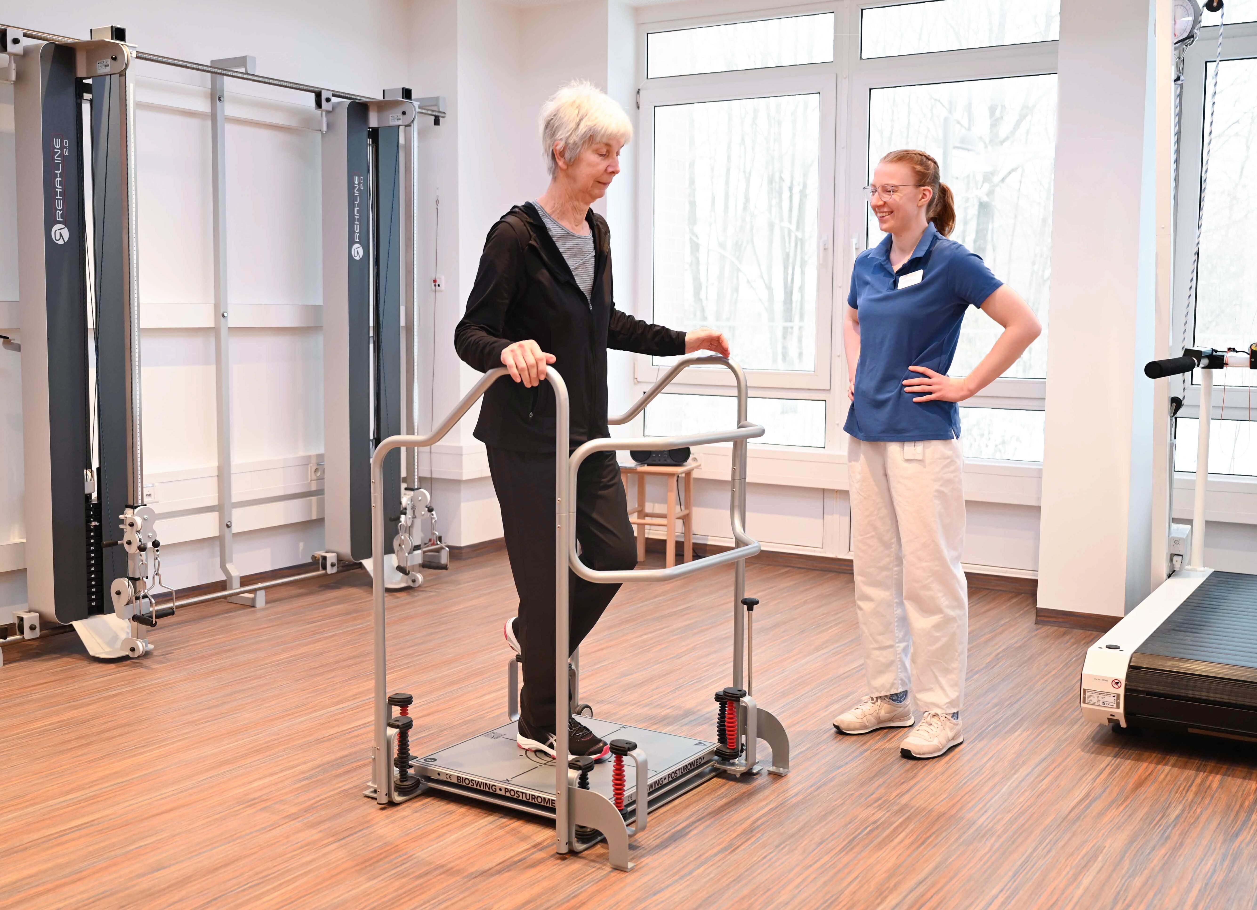 Ältere Dame steht auf einem Gangtrainer wechselnd auf einem Bein ud stützt sich mit beiden Händen an 2 Armstützen. Eine Physiotherapeutin in blauem T-shirt steht neben ihr und begleitet lächelnd.