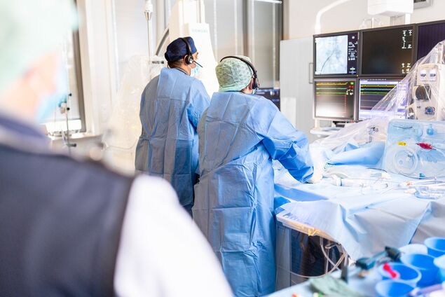 Medizinisch Technische Angestellte steht hinter zwei Operateuren, die konzentriert auf große moderne Monitore blicken