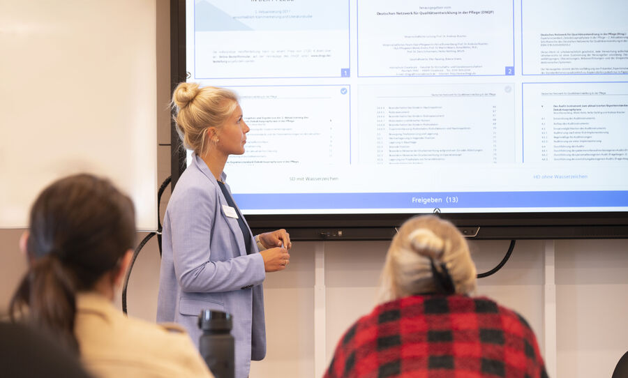 Eine blonde junge Lehrerin steht an einem digitalen Board und blickt auf die Lehrinhalte, während sie diese erläutert..