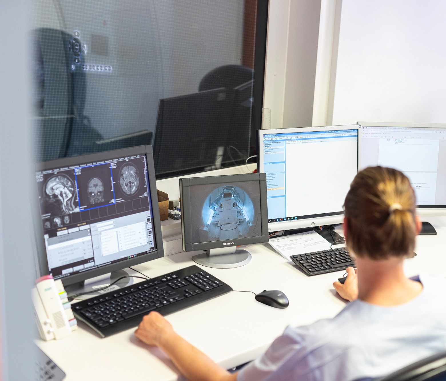 Medizinisch-technische-Angestellte sitzt hinter verdunkelter Scheibe vor einem Monitor mit Patienten Befunden.